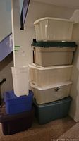 Seven plastic storage bins stacked in basement near staircase and wall.
