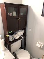 Full view of over-the-toilet cabinet installed above a toilet, showing two doors with glass panels and shelf below with bathroom items
