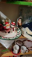 Photo shows holiday platter with holly design, Santa and snowman salt and pepper shakers, and two holiday mugs arranged on a wooden shelving unit.