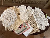 Top view of various handcrafted doilies arranged on a wooden surface including round and rectangular shapes in white and off-white colors, some featuring open crochet work and some dense stitching.