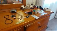 Photo showing multiple jewelry items including necklaces, brooches, earrings, watches, keychains, pins, and a clock on a wooden dresser.