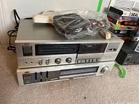 Two stacked vintage JVC audio components: cassette tape player on top with various dials and buttons, receiver below with tuner display and control knobs. Plastic bag with cables and antenna sitting on top.