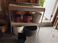 Shelf unit with assembled metal frame and pressboard shelves holding various plastic planters of different sizes and colors, showing weathered condition.