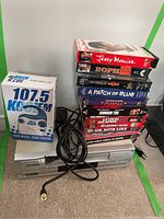 Full view of the lot showing the Sylvania VCR player, remote, KoolFM radio box, and stack of VHS movies on carpet.