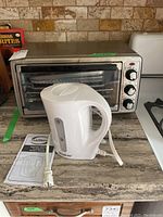 Photo showing the Hamilton Beach toaster oven with three control knobs and the white Simplicite electric kettle with power cord and manual on countertop.