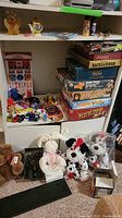 Overview of board games stacked including Rummikub, Backgammon, Yahtzee, puzzles, and scattered small toy cars and stuffed animals below shelf.