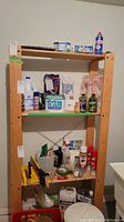 Wood shelf with multiple items on shelves, including food containers, cleaning products, and miscellaneous household items. Shelf is a four-tier natural wood unit with cross-brace support.