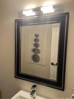 Rectangular mirror mounted on wall above a sink, showing decorative black and silver frame