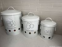 Three white metal containers for kitchen storage labeled for potatoes, onions, and garlic. All have lids and ventilation holes near the base.