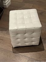 White tufted square ottoman placed on wooden floor with visible stitching and black feet underneath.
