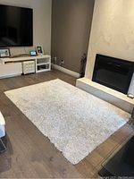 Full view of rectangular white shaggy rug on wooden floor in living room with TV and fireplace.