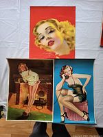 Three vintage pin-up girl calendar prints laid out on fabric: close-up of blonde woman with red background, woman warming by fireplace, woman seated on stool talking on phone in black lingerie.