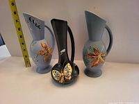 Front view of three Herta pottery vessels showing butterfly and maple leaf designs with measuring tape indicating height.