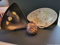 Photo showing 3 pieces of Herta pottery: dark brown triangular dish, grey oval dish with leaves, and brown sugar bowl with lid.