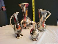 Four metallic lusterware pitchers of varying sizes with reflective surfaces and colorful glaze accents displayed with measuring tape showing largest piece at about 10 inches tall.