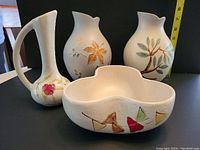 Four pieces of hand decorated Herta pottery displayed together with measuring tape: 2 vases approx 8 inches, vessel with handle 7.5 inches, 3-lobed dish with geometric leaf patterns.
