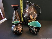 Four pieces of Herta pottery with dark brown glaze and hand-painted autumn leaves design, shown against black background with measurement scale visible showing size of larger pieces