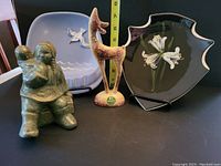 Four hand-decorated Herta pottery pieces shown together: a black Fleur de Lis plaque, blue seagull ashtray, deer figurine, and woman with child figurine.