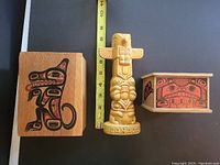 Front view of ceramic totem pole coin bank labeled 'CANADA' with two wooden boxes featuring black and red painted First Nations inspired designs, measured next to a tape ruler.