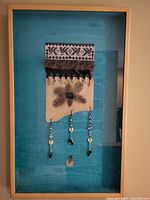 Full view of framed mixed media artwork showing textile, feathers, beads on teal background.
