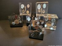 Photo showing all six Lucite cubes with Canadian coins embedded, arranged standing on a black surface, showing multiple coins in each cube.