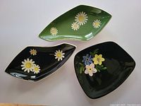 Three hand decorated Herta pottery dishes shown in a group, displaying floral patterns on black and green glazed surfaces.