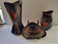 Three pieces of Herta pottery displayed together showing tall vase, medium vase and triangular dish with orange heart lava-style glaze on glossy brown