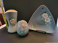 Photo of the three Herta pottery pieces: a triangular plate, tall vase, and smaller oval vase, showing floral hand-painted decoration on blue glaze.