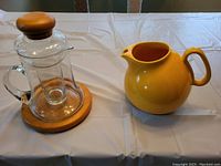 Two pitchers displayed on a white tablecloth: left is a glass pitcher with wooden lid and base, right is a yellow ceramic pitcher.