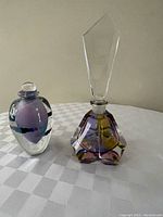 Two crystal perfume bottles shown on a checkered surface, one with a stopper and taller with amber and purple coloring, the other smaller with purple and blue swirls.