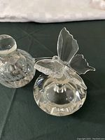 Photo showing two clear crystal perfume bottles with butterfly-shaped applicator tops on a black surface.