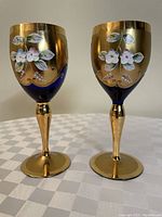 Front view of two vintage Italian wine glasses with gold finish, blue base, and white floral designs.