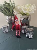 Wide shot showing two reflective metal vases with artificial greenery and flowers, three winter tree designed glass votive holders, and a red/white elf doll.