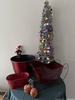 Full view of the lot showing the silver tinsel collapsible 3-foot Christmas tree with colorful round ornaments, the large red metal flower pot, small elf figurine, two small decorative pumpkins, the smaller red flower pot, and the red metal decorative sleigh.