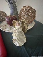 White ceramic angel figure seated with gold wings on a decorative plate, next to a wicker twig sphere and burgundy apple-shaped candle holder.