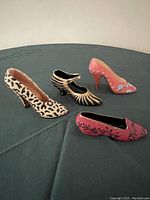 Four miniature resin high heel shoes displayed on a dark green cloth, showing various painted decorative patterns: leopard print, zebra stripes, coral floral, and red floral.