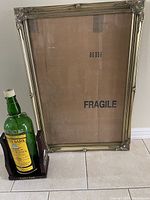 Full view of green Cutty Sark whiskey bottle mounted on wooden swivel stand placed beside ornate gold-tone picture frame with protective backing labeled 'Fragile'.