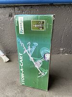 Photo of the Stow-A-Cart golf pull cart in its original green packaging showing cart folded with wheels and handle visible.