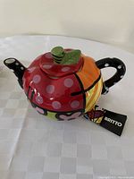 Colorful Romero Britto apple-shaped tea pot with red polka dot body and green leaf lid.