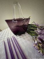 Purple glass tote-style vase with clear glass handles and a bunch of faux purple wisteria flowers, six tapered purple candles grouped beside it on a white checkered tablecloth.