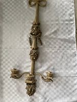 Full view of the ornate gold-toned wall sconce with three candle holders lying flat on a white textured surface. Shows detailed decorative motifs and overall shape.