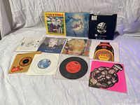 Photo showing 12 vinyl 45 RPM singles in paper and original graphic sleeves laid out on a white cloth background.