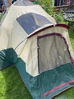 Full view of beige and green 4 person Woods tent set up on grass with entrance and covered vestibule visible, mesh window on front flap.