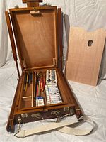 Portable wooden artist easel folded open showing storage compartments with art supplies inside and removable wooden palette leaning to the side.