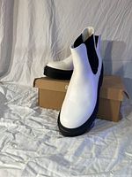 White Cushionaire boots shown standing on Cushionaire branded box on fabric backdrop from front angle, showing black thick outsole and black elastic side panels.