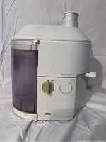 Front and side view of white Braun juicer showing green on/off dial, transparent purple pulp container, and juice spout.