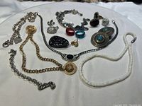 Photo showing a variety of costume jewelry including a gold medallion necklace, silver chains, white shell bead necklace, blue rings, and dark carved brooch.