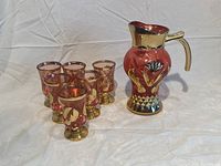 Full set showing a red glass pitcher with gold painted floral and geometric decoration, and six matching red glasses with gold designs.