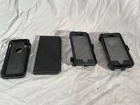 Three OtterBox iPhone holders laid out on a white cloth background: one wallet style phone case with magnetic back and leather-like finish, two rugged cases with black rubber edges and clear front covers.