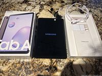 Samsung Galaxy Tab A box, tablet powered on showing Samsung screen, and charging cable with power adapter in the box tray.